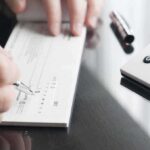 The Shocking Downside of Lending to Friends Person writing a check on a reflective surface with a calculator nearby