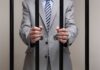 Anti-Corruption Hero BUSTED—Massive Bribe Bombshell Businessman in a suit holding prison bars with handcuffs on his wrists