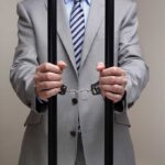 Businessman in a suit holding prison bars with handcuffs on his wrists