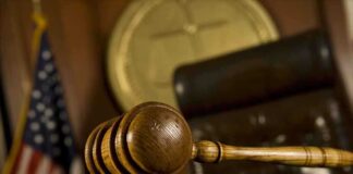 A wooden gavel resting on a desk in a courtroom with an American flag in the background