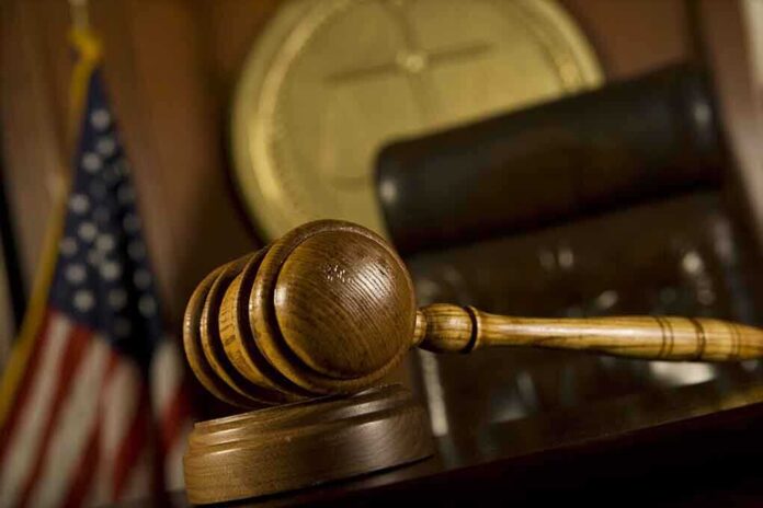 A wooden gavel resting on a desk in a courtroom with an American flag in the background