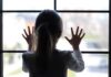 A child with long hair presses their hands against a window, looking outside
