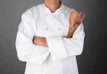A chef in a white uniform holding wooden cooking utensils with crossed arms