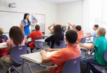 Teacher in a blue dress instructing students in a classroom with hands raised