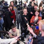 A crowded press conference with journalists and cameras surrounding a speaker
