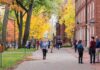Students walking on a campus path surrounded by autumn trees and brick buildings