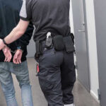Crypto Plot To Attack LA Jewish Center EXPOSED Officer escorting handcuffed person down hallway.