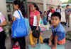 People waiting outside carrying bags, boys sitting and playing.