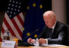 Biden’s Audio Battle: What’s He Hiding? Man seated at table with flags in background.