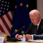 Biden’s Audio Battle: What’s He Hiding? Man seated at table with flags in background.