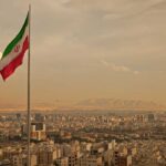 Trump’s Iran Strike PAUSED: Deal Or Deception? Iranian flag waving over a city skyline with mountains in the background
