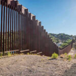 Border Wall Ultimatum Hits Texas Tall metal border wall with rural landscape.