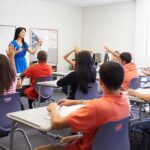 School Demographics Flip — Guess Who Shrank Teacher in a blue dress instructing students in a classroom with hands raised