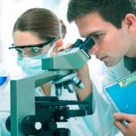 Child’s ‘Fatal’ Tumor Vanishes — Stuns Doctors Two scientists conducting an experiment in a laboratory, one looking through a microscope and the other holding a flask with blue liquid