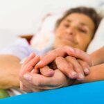 Death Rate Tumbles—The Twist Nobody Expected A caregiver holding the hands of an elderly patient in a hospital bed