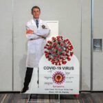 Fauci Crisis After COVID Vaccine Ignites Washington Cardboard cutout of doctor with COVID-19 virus info display