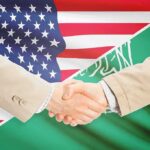 Handshake between two people over U.S. and Saudi flags