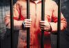 Person in an orange jumpsuit gripping prison bars in a dark cell