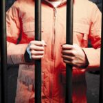 Judge Sparks Fury After Triple-Fatal Crash Person in an orange jumpsuit gripping prison bars in a dark cell