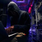 Hooded person using a laptop in a dark server room