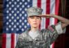 soldier in camouflage uniform saluting in front of a U.S. flag