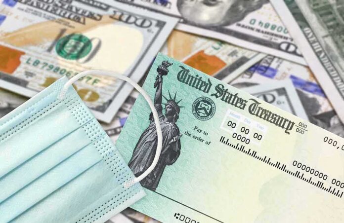 shutterstock_1935630070 U.S. Treasury check with face mask on pile of hundred-dollar bills