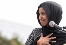 Feds Hit Rep. Ilhan Omar’s Home – What Did They Find? Ilhan Omar speaking at a microphone with hand on chest